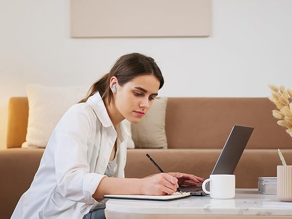 Woman wearing wireless earbuds works on laptop at desk with notebook and coffee mug. Home office setting with sofa and decor visible in background.