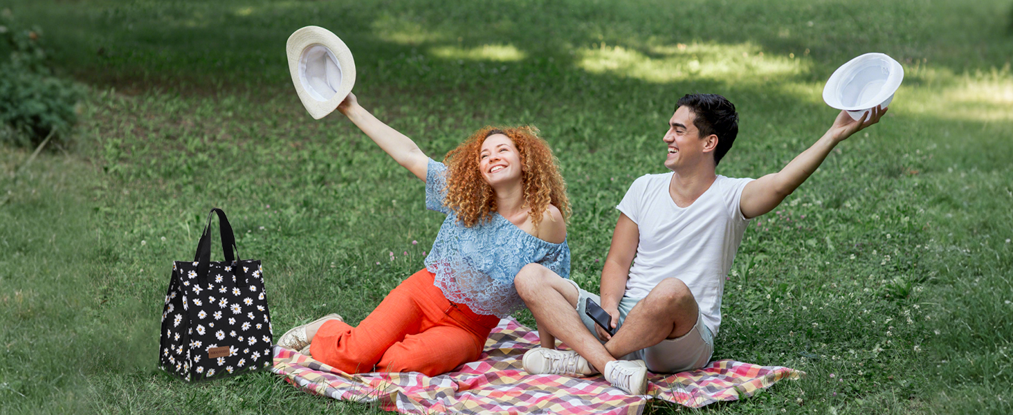 Outdoor picnic scene on grass with blanket and polka dot bag. Two people sitting casually outdoors, holding up white sun hats while enjoying a sunny day.