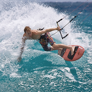Kitesurfer performing aerial maneuver over turquoise ocean water, holding control bar while airborne above red and white kiteboard during active water sport demonstration.
