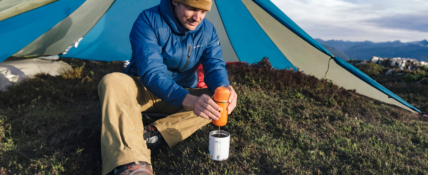 Person in blue jacket sitting outside tent, holding orange water bottle. Camping scene with mountainous background.