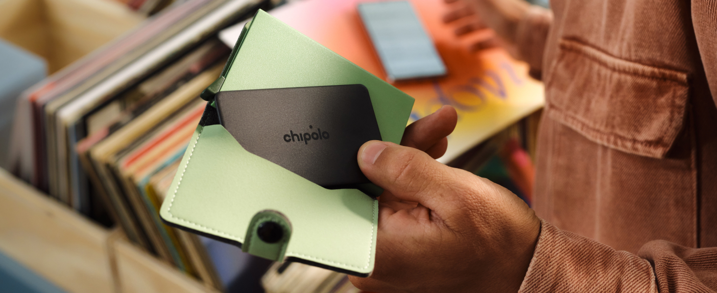 Close-up of hands holding black rectangular portable speaker or audio device with yellow and green accent colors visible in blurred background with books.