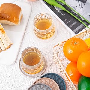 Lifestyle product photo featuring citrus fruits, glass tumblers with amber liquid, and decorative coasters with embossed geometric patterns.