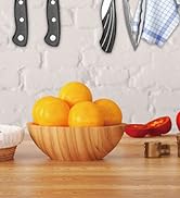 Kitchen scene with wooden bowl filled with oranges. Visible are kitchen utensils, a blue checkered cloth, and partial views of other ingredients.