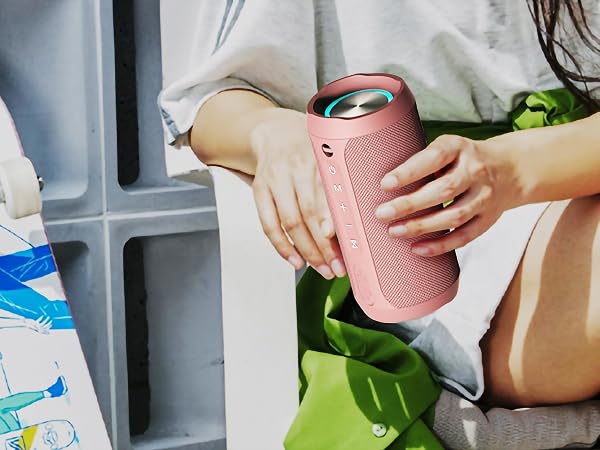 Hands holding a cylindrical pink portable Bluetooth speaker with a textured surface and visible control buttons. The speaker is being carried alongside colorful beach items.