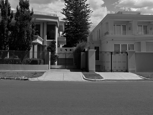 Black and white image of residential street with large houses, fences, and trees. Two-story homes with pillared entrances visible.