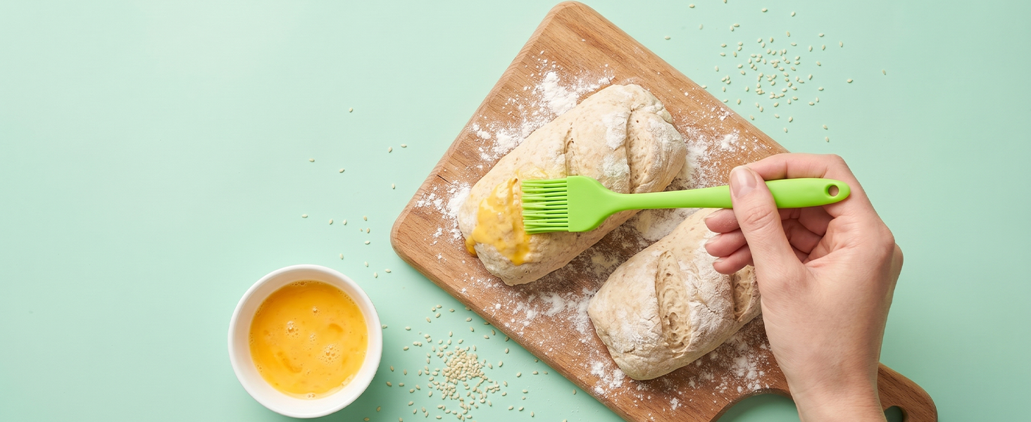Green silicone brush applying egg wash on homemade bread dough with sesame on mint background