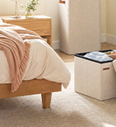 Beige upholstered bed with light pink/blush bedding and pillows, positioned against a light wooden headboard in a minimalist bedroom setting.