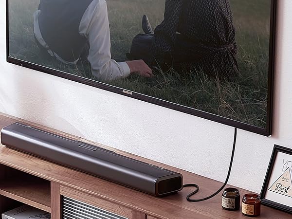 Long, slim soundbar beneath a wall-mounted TV displaying a nature scene. Soundbar sits on wooden TV stand with shelves. Small jars and framed picture visible.