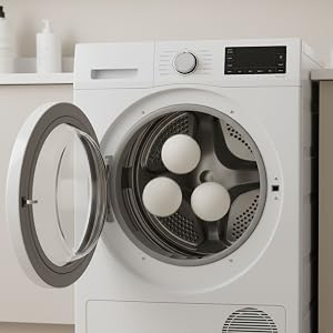 Front-loading dryer with open door showing three white dryer balls inside the stainless steel drum. Digital display and control dial visible on white appliance exterior.