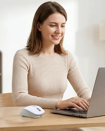 Woman with long brown hair wearing beige ribbed knit sweater typing on laptop with white mouse on wooden desk in bright home office interior with plant and lamp.