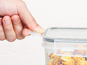 Clear plastic food storage container with airtight lid being sealed by a finger pressing on the corner, demonstrating its locking mechanism.