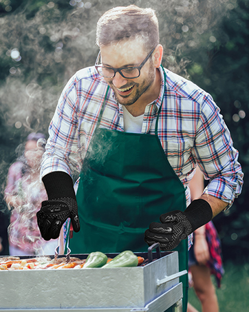 Person in plaid shirt and green apron grilling outdoors. Wearing black gloves, tending to food on a smoking grill with vegetables visible nearby.