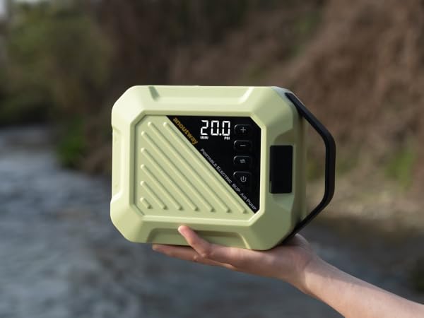 Hand holding a light green portable electronic device with digital display showing '20.0'. Device has rugged design, diagonal striped pattern, and appears to be used outdoors near water.
