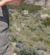Outdoor nature scene showing blurred purple flowering plants or wildflowers in background, person's legs in denim pants visible in soft focus.