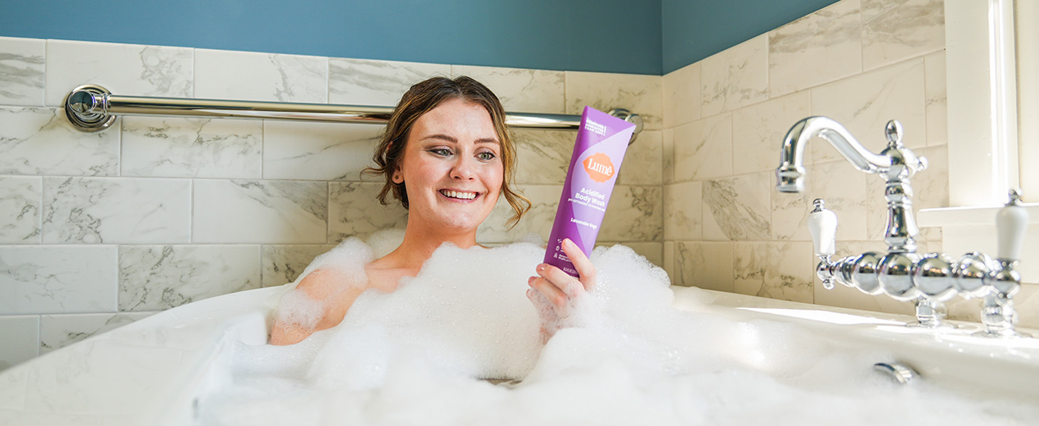 A woman using Lume Acidified Body Wash in Lavender Sage being used in a bath tub