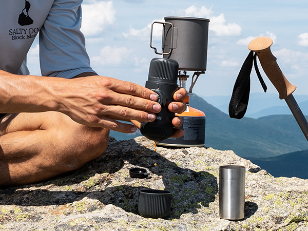 Portable camping stove and cookware being assembled on rocky mountain terrain, with scenic mountain vista in background. Multiple metal cooking components visible.