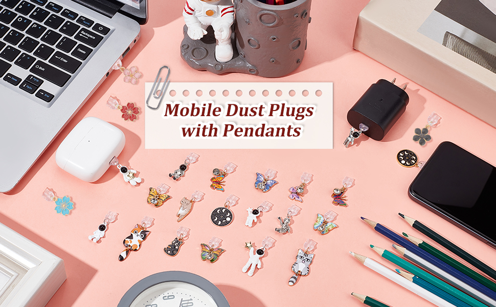 Collection of decorative mobile phone dust plugs with pendant designs displayed on pink surface alongside electronics and stationery items.