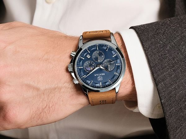 Wristwatch with blue dial worn on person's wrist wearing white dress shirt and brown leather strap. Chronograph-style watch face with subdials visible on male wrist.
