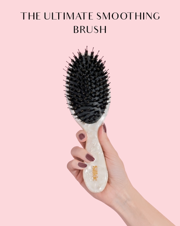 Oval-shaped hair smoothing brush with black bristles and white handle, held in a hand against a pink background. Text reads 'THE ULTIMATE SMOOTHING BRUSH'.
