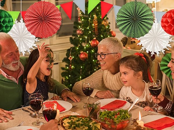 Family gathered around Christmas dinner table with festive decorations including colorful paper fans and a decorated tree in the background.