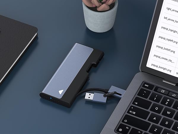 Sleek portable SSD drive in gray, connected to laptop via USB cable. Surrounded by notebook, pen, and papers on dark surface.
