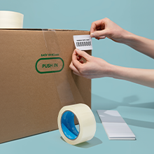 Clear package tape sealing a cardboard box and a pair of hands applying a direct thermal label