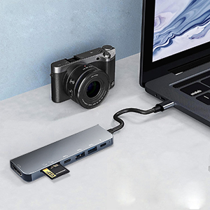 Laptop connected to compact USB-C hub with multiple ports. Digital camera and memory card nearby on light surface.