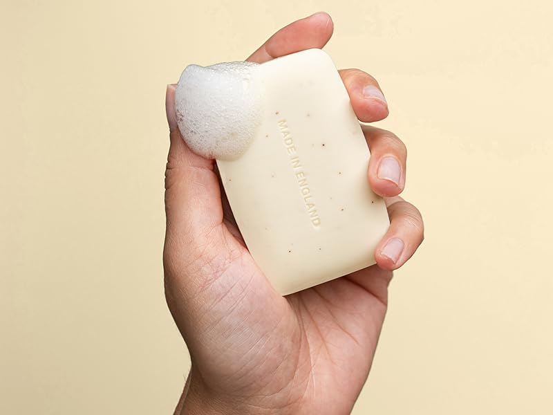 Cream-colored rectangular bar soap held in a human hand against beige background. Product shows smooth texture with subtle speckled pattern throughout.