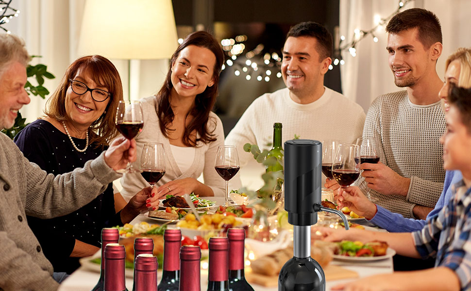 Group gathering around a table with wine glasses, food, and an electric wine opener in the foreground. Festive atmosphere with string lights visible.