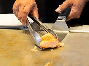 Hands using a metal kitchen tool to scoop or cut a round, orange food item on a wooden surface.