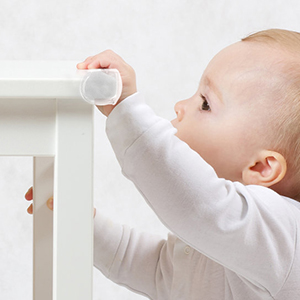 Baby in white clothing reaching up to touch a round, white object on a table edge or surface.