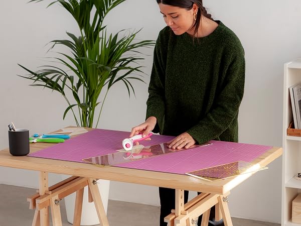 Person at wooden trestle table with purple cutting mat, using crafting tools with indoor plant in background.