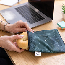 Woman's hands removing a bagel from a dark colored cloth snack bag on a desk next to a laprtop