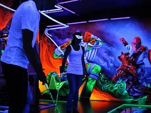 Colorful mini-golf course with neon-lit obstacles and figures in a dimly lit indoor space. People playing amid vibrant green, blue, and red elements.
