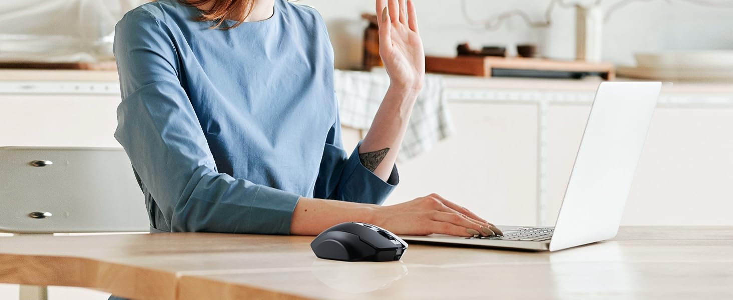 Person in blue shirt sitting at desk with laptop, holding hand up as if waving or gesturing during video call.