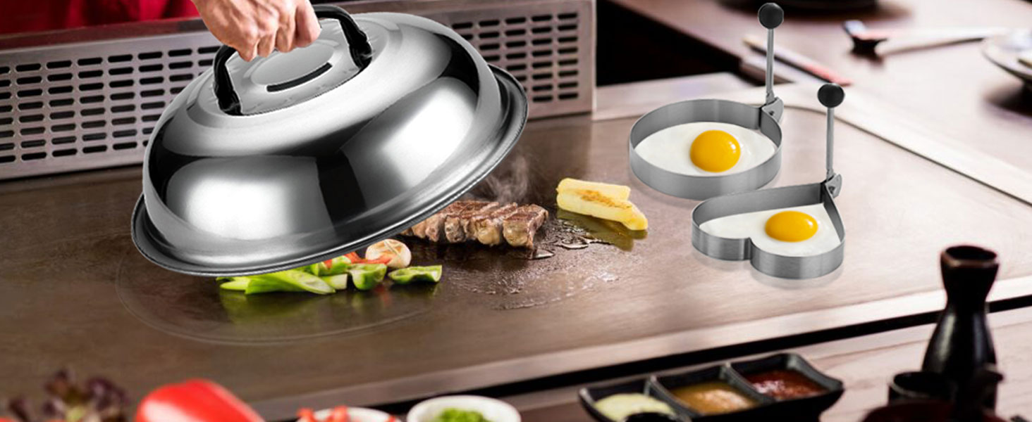 Stainless steel cooking dome lifted over grilled meat and vegetables. Nearby are egg-shaped molds with fried eggs on a wooden surface.