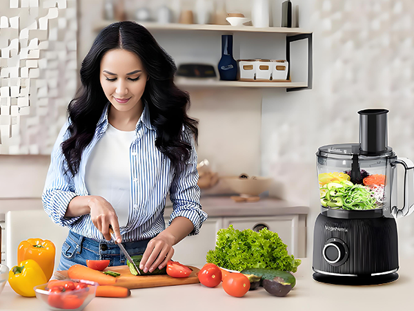 Kitchen scene with food processor on counter containing fresh vegetables. Fresh produce and orange juice visible on cutting board.
