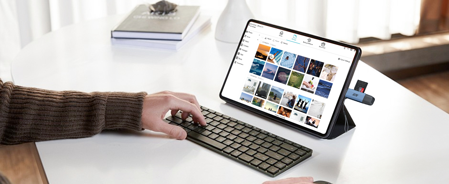 Tablet device propped up with wireless keyboard on white desk surface, showing grid of app icons on screen. Work setup in home environment with natural lighting.