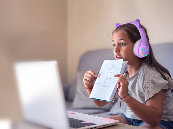 Child wearing purple cat-ear headphones, looking at laptop screen while holding a notebook. The headphones feature illuminated ears and plush ear cushions.