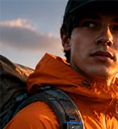 Portrait of person wearing orange and black outdoor jacket against cloudy sky background. No product visible; appears to be lifestyle or brand image.