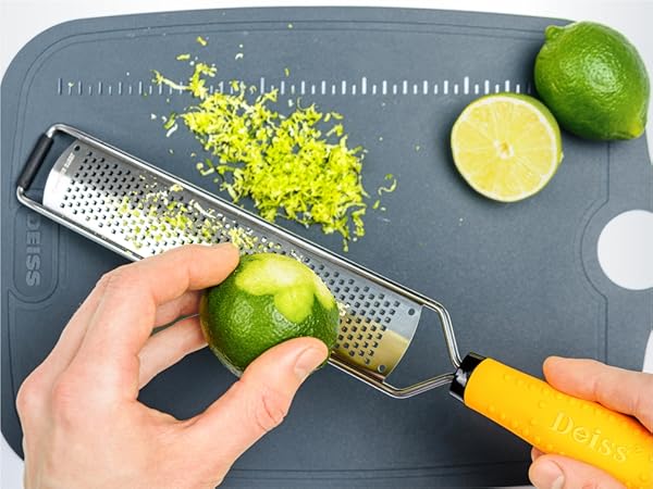 Hand grating lime zest with a yellow-handled grater on a gray cutting board. Whole and halved limes visible.
