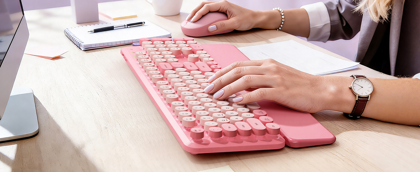 Woman typing on a Blush Pink retro wireless keyboard with matching mouse on a bright office desk.