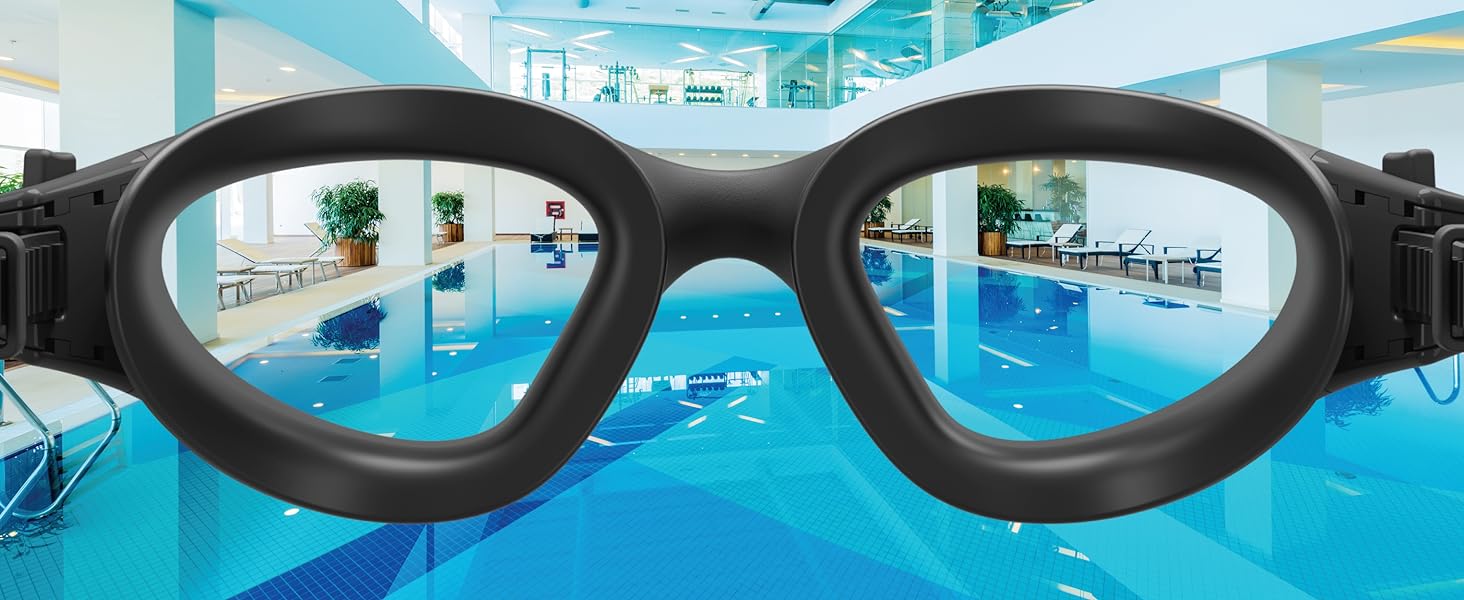 Black swimming goggles with wide frames, showing a view through the lenses of an indoor swimming pool with blue water and lounge chairs.