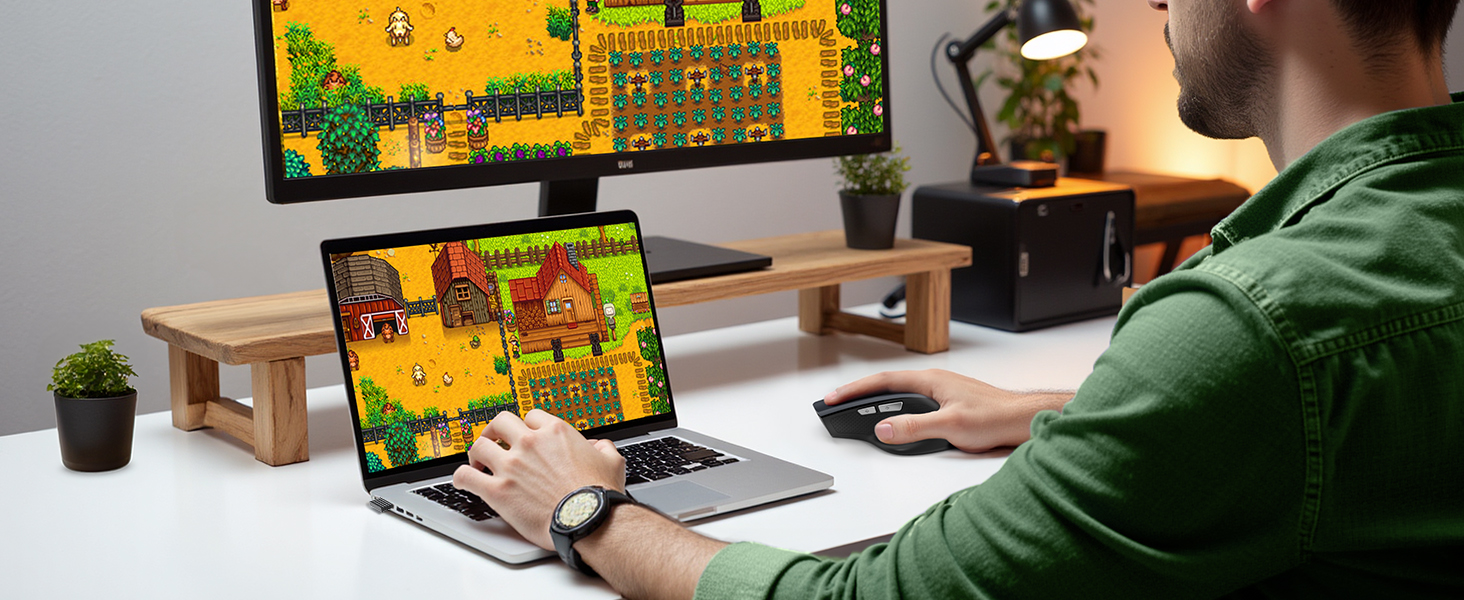 Person using laptop displaying colorful pixelated game. Second monitor shows same game. Desk setup includes wooden stand, plants, and smartwatch on user's wrist.
