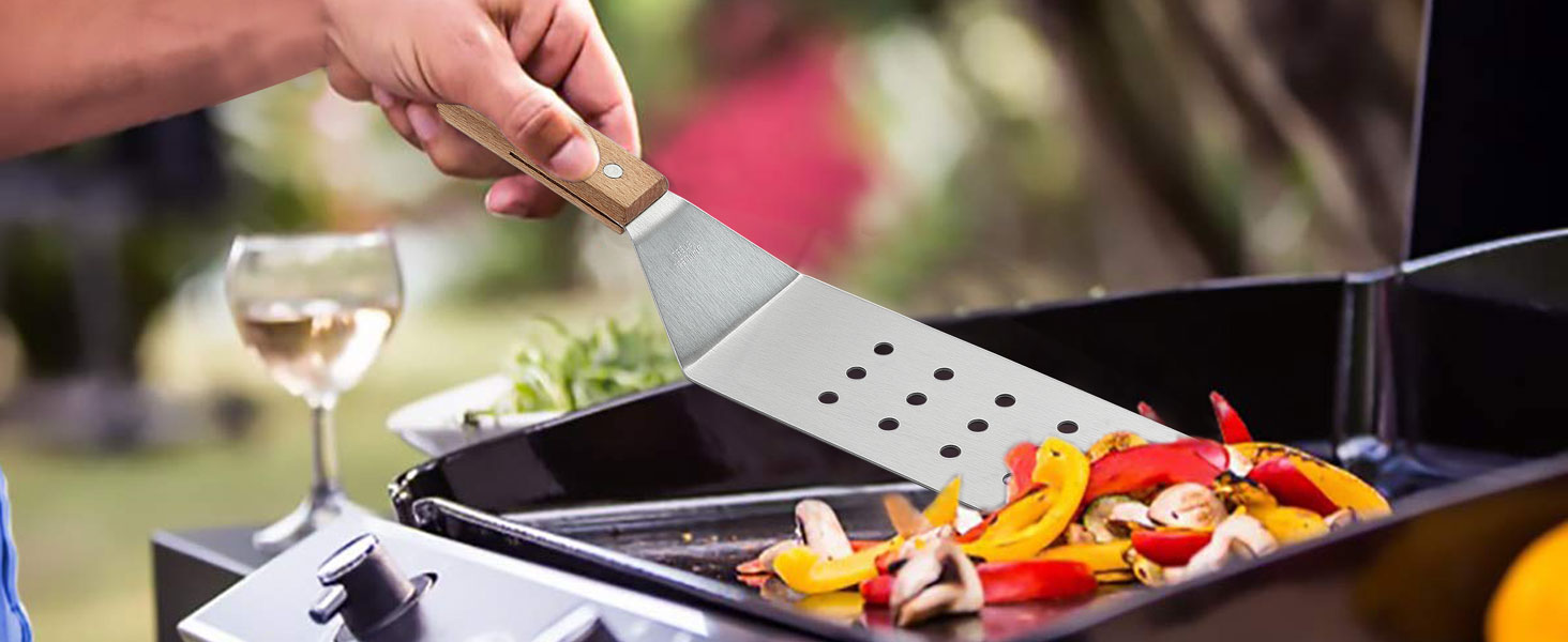 Hand using metal spatula with holes to cook colorful bell peppers and vegetables in a pan. Wine glass visible in background.