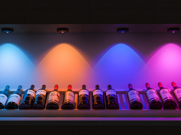 Row of wine bottles on a shelf, illuminated by colorful spotlights in blue, orange, purple, and pink hues.
