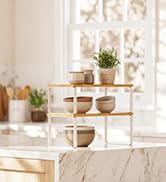 Bright interior room with white wall décor, pendant lights, wooden furniture, potted plants, and decorative objects arranged on shelves and surfaces.