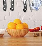 Kitchen scene with wooden bowl filled with oranges. Visible are kitchen utensils, a blue checkered cloth, and partial views of other ingredients.