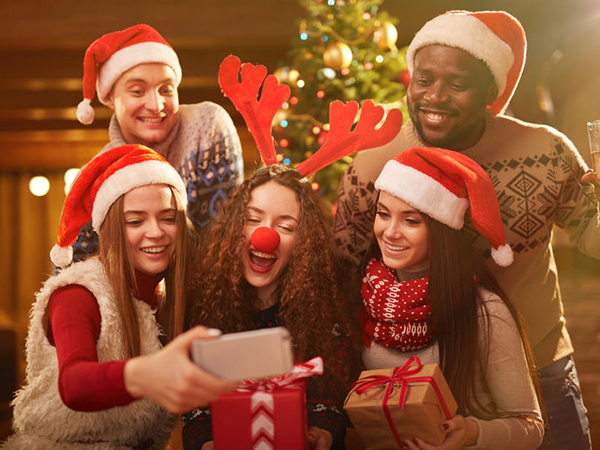 Group of people wearing Santa hats and holiday sweaters, gathered together taking a selfie. Christmas tree visible in background.