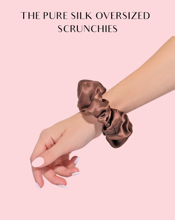 Oversized silk hair scrunchie in brown, worn on a wrist against a pink background. Text reads 'THE PURE SILK OVERSIZED SCRUNCHIES'.